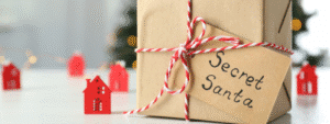 A kraft paper wrapped gift box tied with red and white striped string and a handwritten tag reading "Secret Santa," positioned in front of a blurred festive background featuring miniature red house decorations and twinkling lights. The presented package captures the anticipation and fun of exchanging budget-friendly secret Santa gift ideas during the holiday season.
