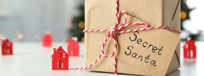 A kraft paper wrapped gift box tied with red and white striped string and a handwritten tag reading "Secret Santa," positioned in front of a blurred festive background featuring miniature red house decorations and twinkling lights. The presented package captures the anticipation and fun of exchanging budget-friendly secret Santa gift ideas during the holiday season.