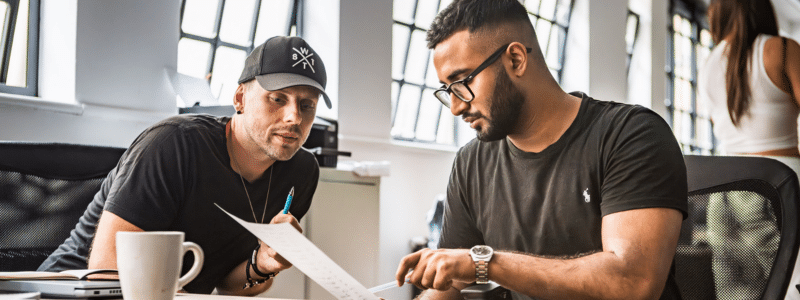 Two men sitting at a table in a bright office space, one wearing a cap and the other wearing glasses, reviewing documents together, illustrating the process of discussing and understanding what is a guarantor and the responsibilities involved in rental agreements.