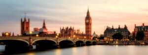 A panoramic view of London's Westminster skyline at dusk, featuring the iconic Big Ben clock tower and Houses of Parliament illuminated against a pastel pink and blue twilight sky, with Westminster Bridge spanning the Thames River in the foreground and historic buildings creating a dramatic silhouette along the waterfront.