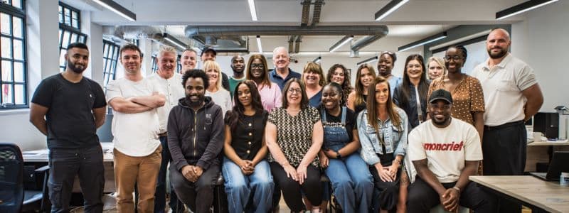 A diverse and friendly team of approximately 20 professionals from Housing Hand standing together in a modern, bright office space with industrial-style windows and exposed ductwork. The welcoming group displays an approachable and professional atmosphere, representing the experienced and trustworthy team behind Housing Hand that landlords, letting agents, and accommodation providers work with when choosing a guarantor partner to provide reliable financial protection and support for rental agreements.