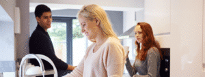 A blonde woman in a beige knit sweater stands at a modern kitchen sink looking downward, while two housemates interact in the background - a man in a dark sweater and a red-haired woman in a grey top holding a cup of tea. The bright, contemporary kitchen with white cabinets and large windows creates a welcoming atmosphere that captures the social dynamics and everyday moments of living in a shared house