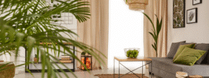 A bright, inviting living room viewed through lush green palm fronds in the foreground, featuring a grey sofa with olive green cushions, a wooden coffee table with a decorative bowl, beige curtains, a woven pendant light, botanical wall art, and natural textures throughout. The warm, personalized space demonstrates thoughtful styling and homey touches that illustrate practical ways of making your rental property feel like a home.